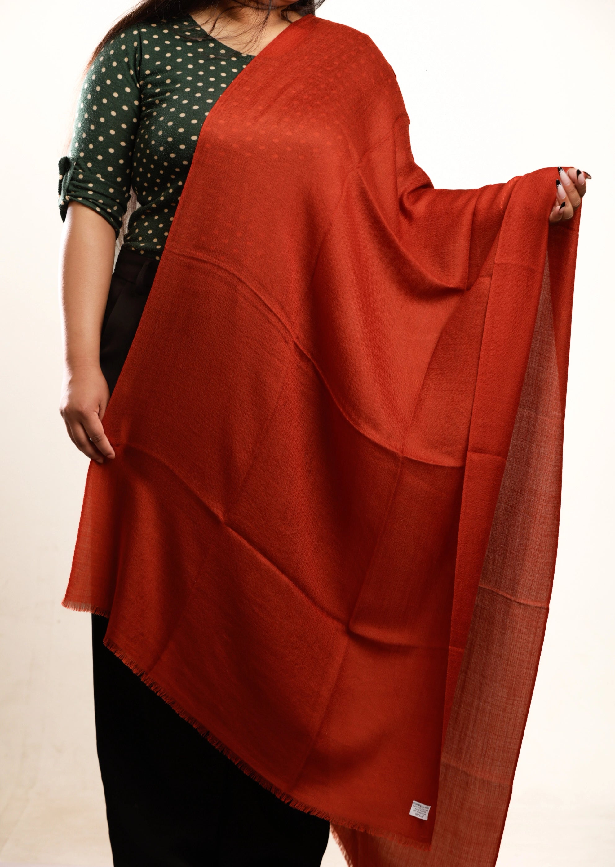 A person wearing a dark green and white polka dot top and black pants, holding up a large rust-orange shawl or scarf with a subtle textured border.