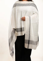 A person wearing a large cream and gray striped shawl or wrap draped over the shoulders, paired with black pants, against a plain light background.
